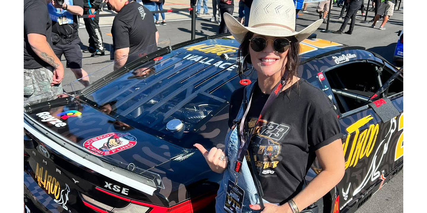 Jenna Paulette, con un sombrero de vaquero de paja, está detrás de un auto de carreras negro número 23 que muestra el logotipo rojo, blanco y negro de Justin Cowboy Crisis Fund.